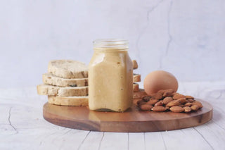 A jar of creamy almond spread on a wooden board with scattered almonds and slices of breads